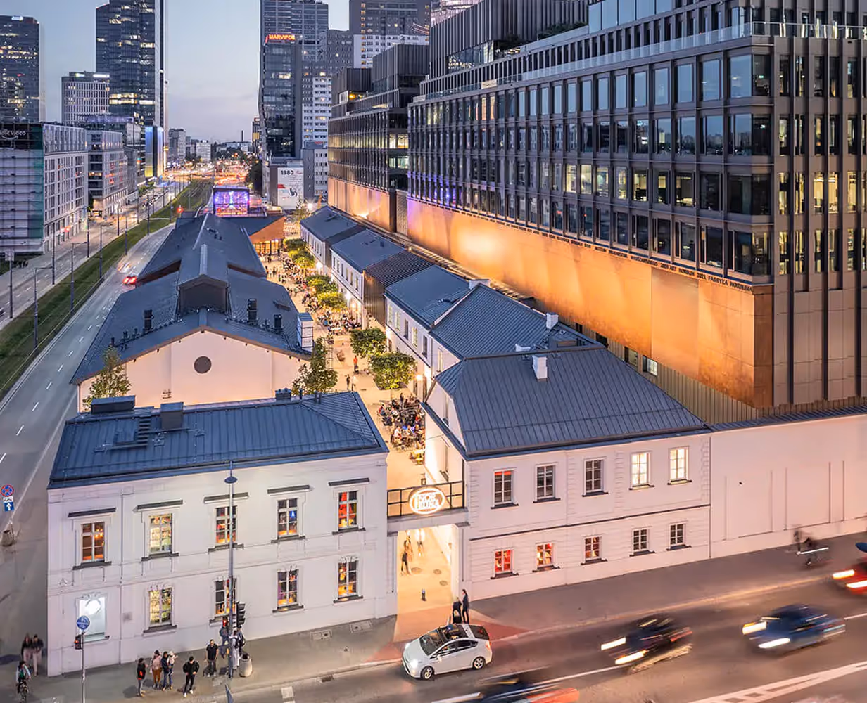 Wieczorny widok na odnowiony kompleks budynków z białymi ścianami i szarymi dachami w centrum miasta z ruchem samochodowym i nowoczesnymi wieżowcami w tle.