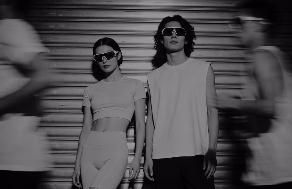 Para w sportowych ubraniach i okularach przeciwsłonecznych stoi przed metalową roletą, podczas gdy dwie rozmazane postacie biegną obok nich.