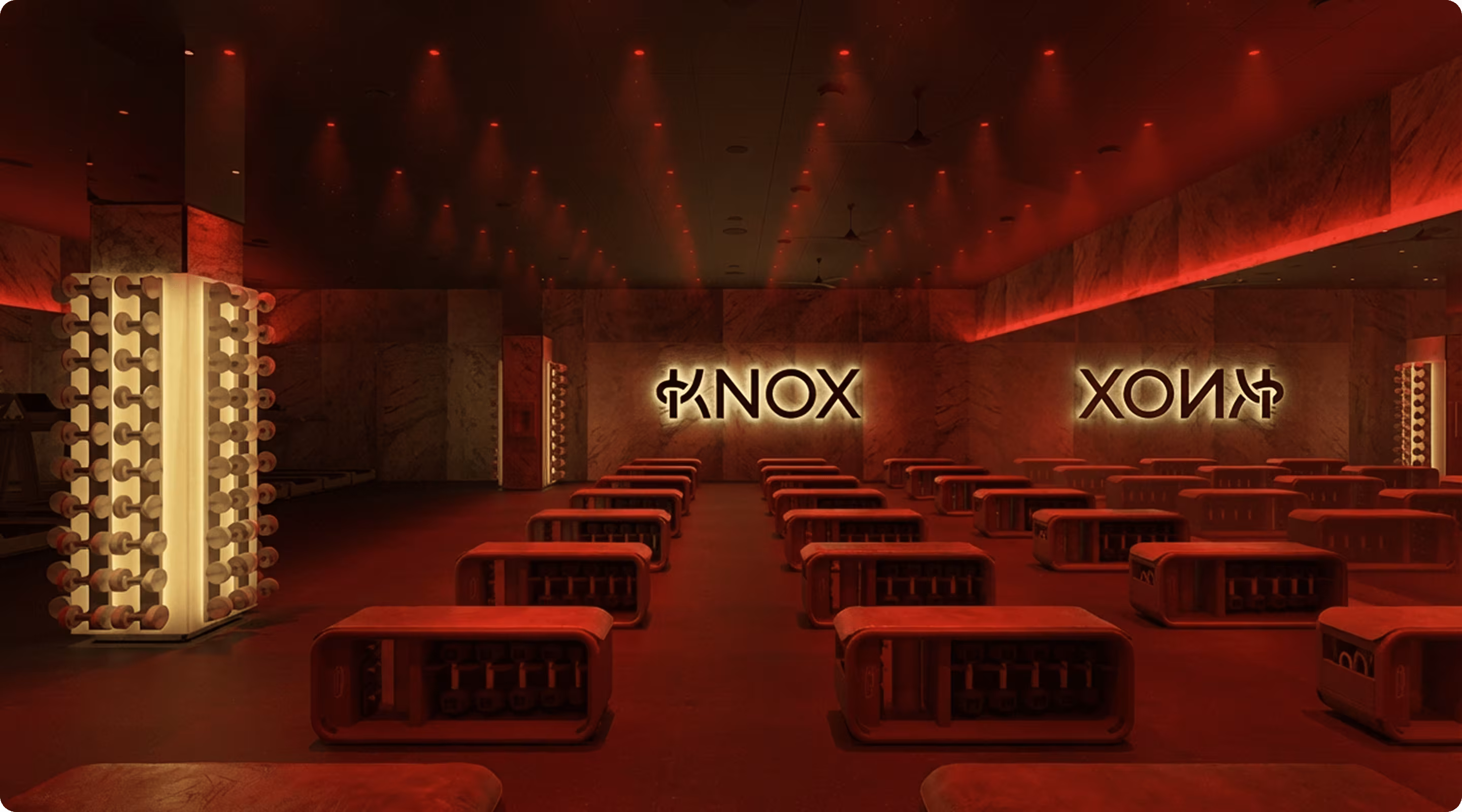 Nowoczesna siłownia z ławkami i sztangami o czerwonym oświetleniu oraz podświetlanymi znakami KNOX na ścianie.