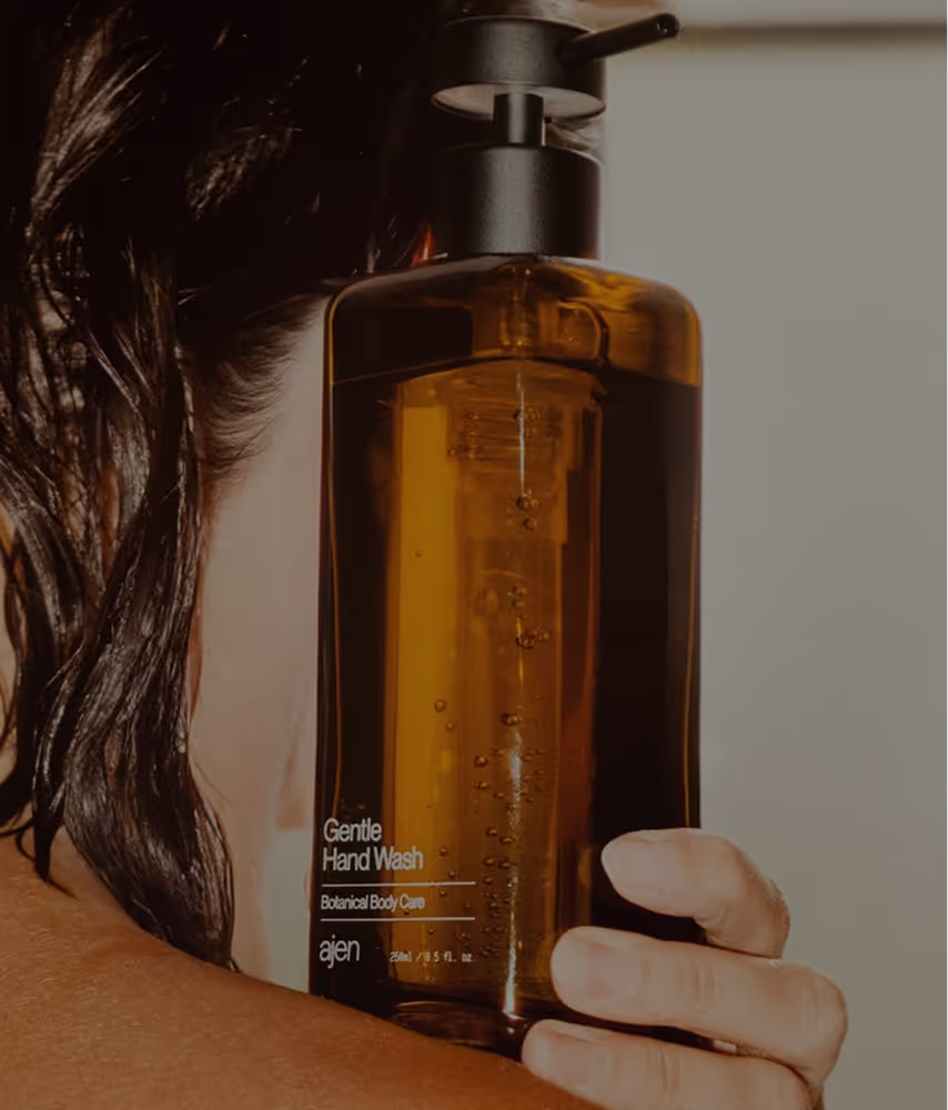 Butelka łagodnego mydła do rąk Botanical Body Care marki ajen trzymana przy ramieniu osoby o wilgotnych ciemnych włosach.