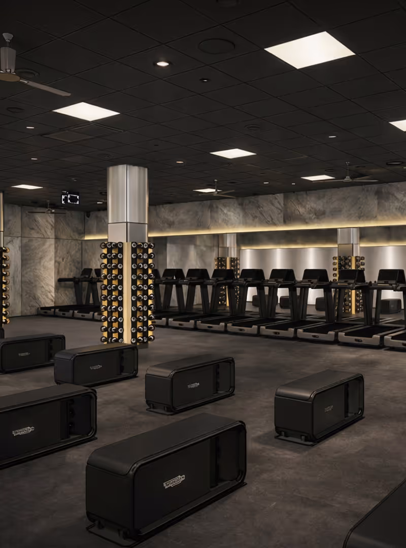 Elegancka siłownia z czarnymi ławkami do ćwiczeń, bieżniami przy ścianie oraz stojakami na hantle przy kolumnach i lustrze.