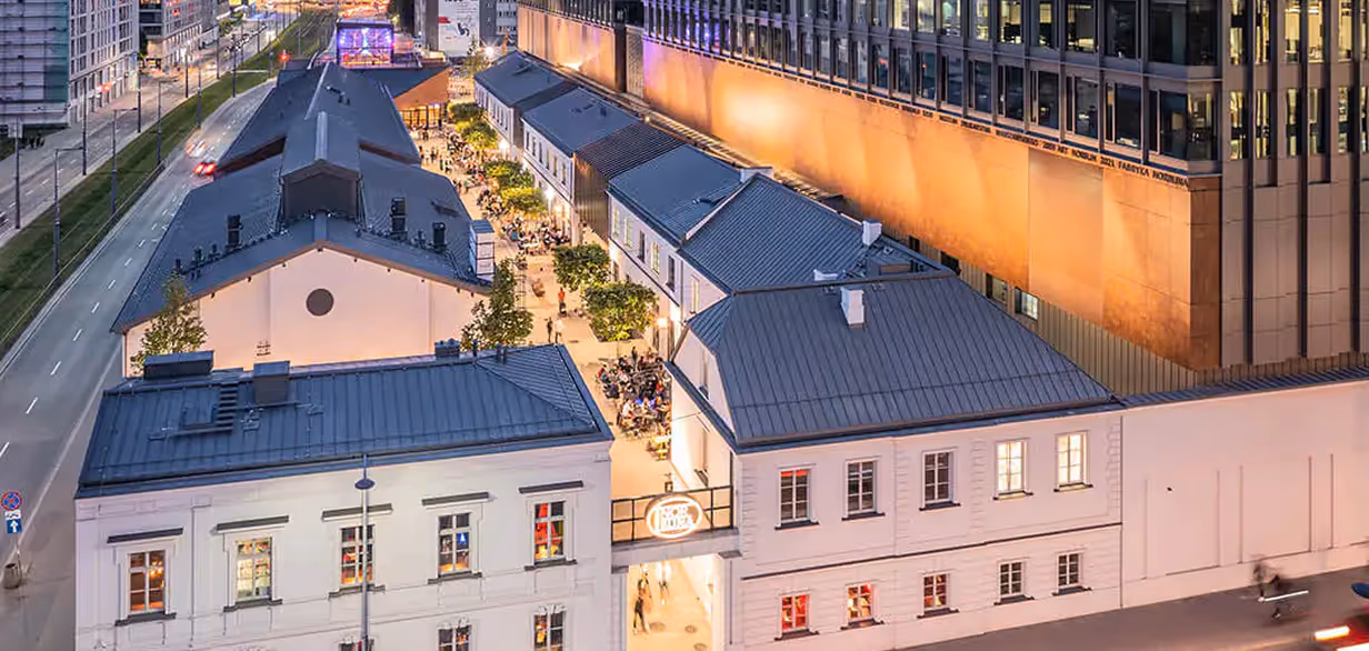 Widok z lotu ptaka na oświetloną ulicę z białymi budynkami i ludźmi siedzącymi przy stolikach na zewnątrz wieczorem.