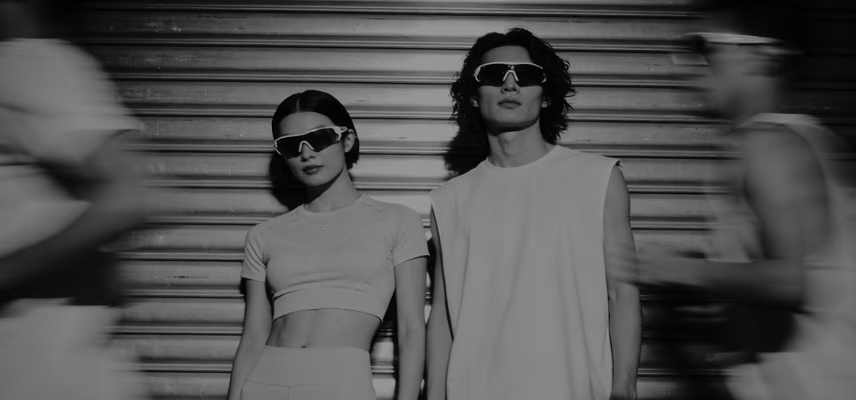 Dwoje młodych ludzi w sportowych ubraniach i okularach przeciwsłonecznych stoi nieruchomo na tle metalowej żaluzji, podczas gdy inni ludzie mijają ich w ruchu.