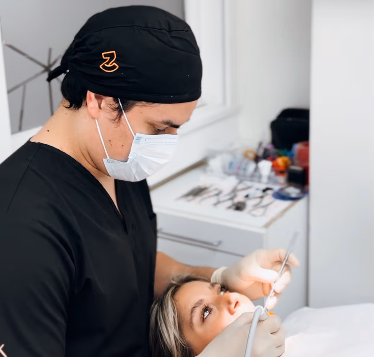 Dentista con mascarilla y gorro negro atendiendo a paciente reclinada en sillón dental en consultorio.