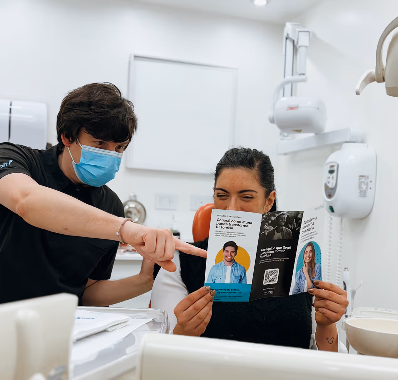 Un dentista con mascarilla señala un folleto que sostiene una paciente sentada en una clínica dental.