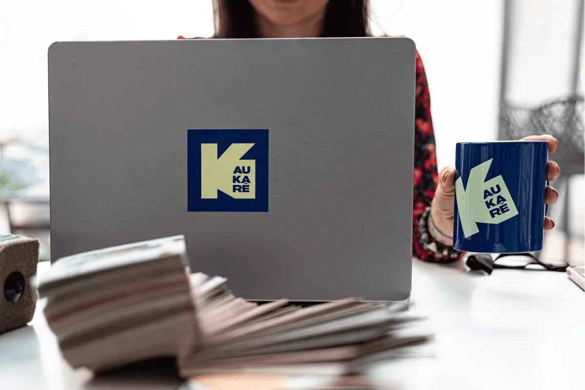 Personne tenant une tasse bleue avec un logo "K AUKARÉ" devant un ordinateur portable équipé du même logo sur un bureau.
