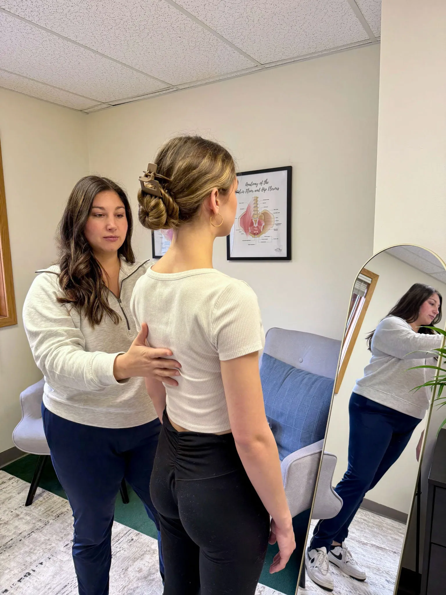 A woman in white and navy blue assessing the posture of another woman wearing a white top and black leggings in a room with a mirror and an anatomy poster on the wall.