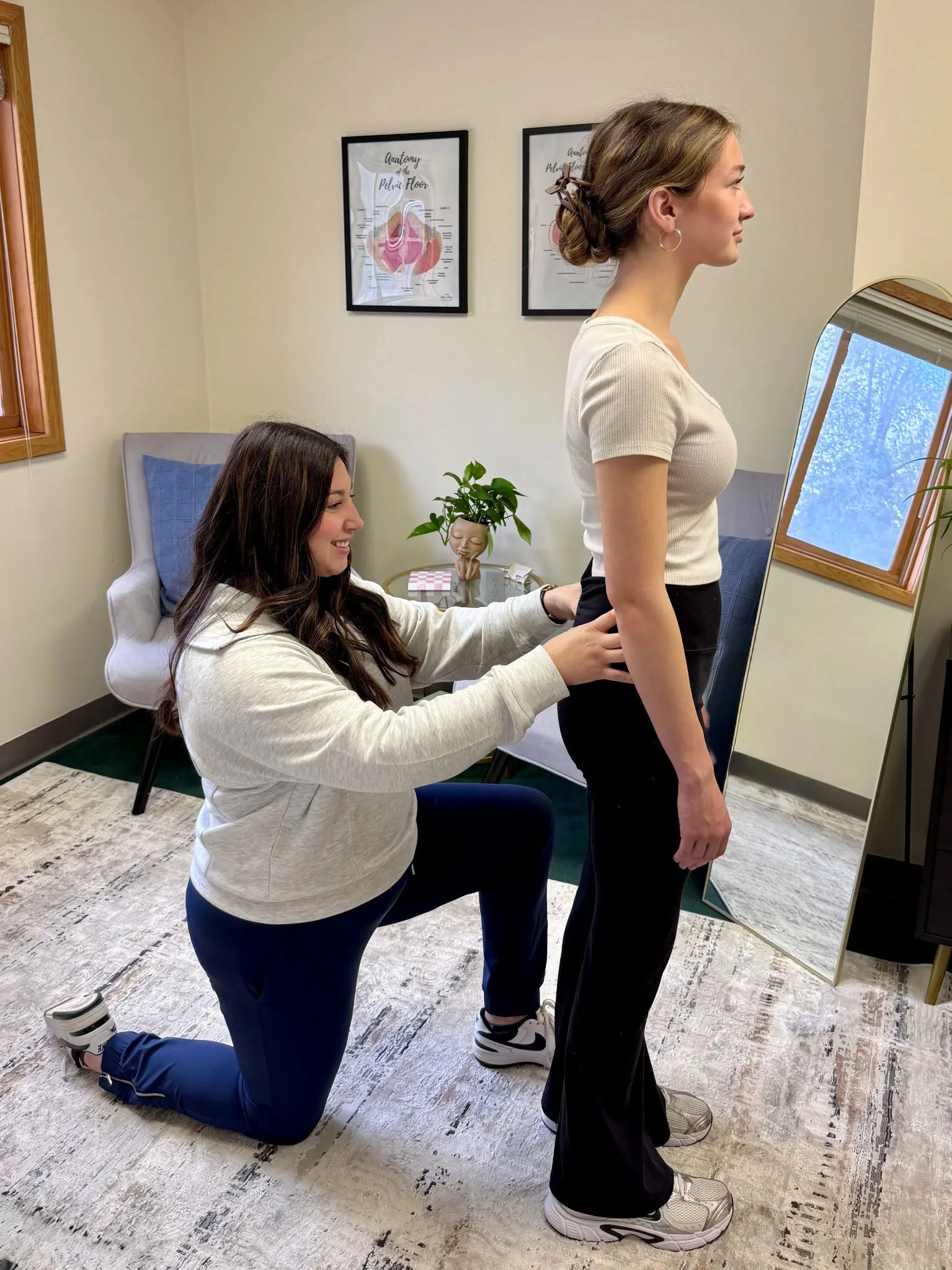 A woman kneeling adjusts the posture of another standing woman in a room with anatomy posters and a mirror.