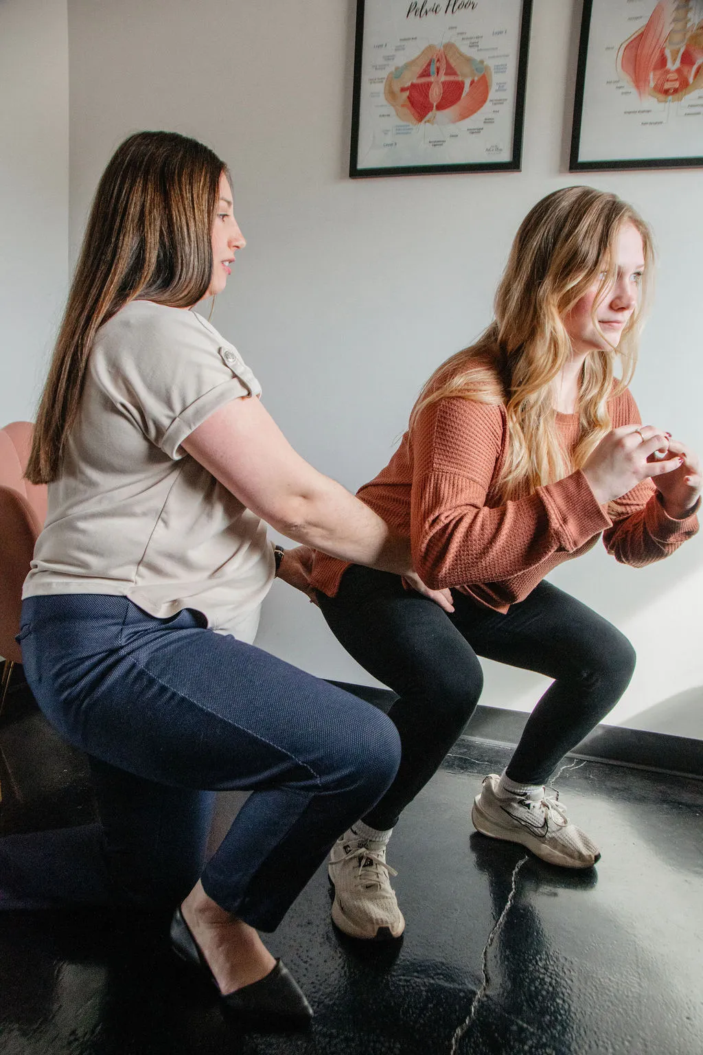 A woman assisting another woman in a squatting exercise indoors with pelvic floor anatomy charts on the wall.