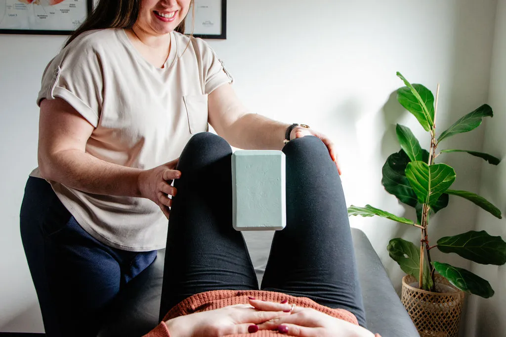 A therapist guiding a client lying on a table with a yoga block between their knees in a bright room with a potted plant nearby.