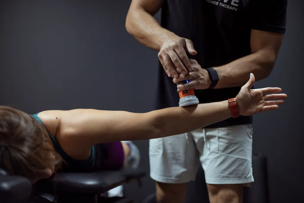 Close-up of a therapist using a handheld massage device on a woman's extended arm during a physical therapy session.