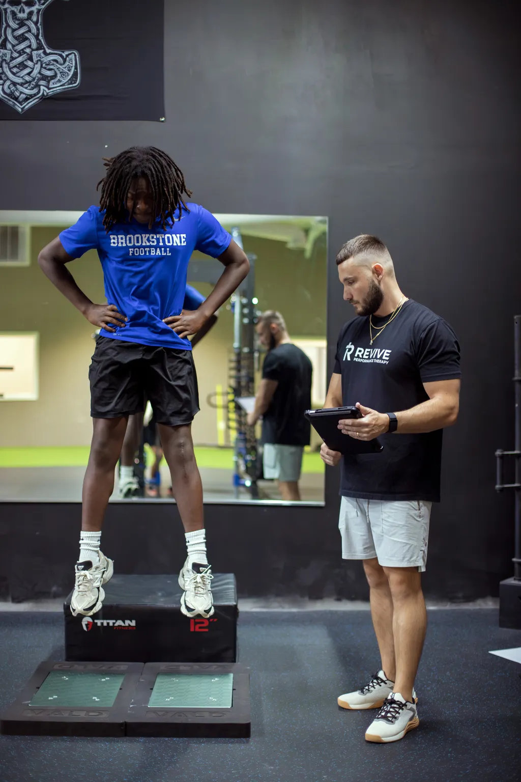 Young athlete in blue Brookstone Football shirt standing on a 12-inch Titan Fitness platform while trainer in black Revive Performance Therapy shirt records data on a tablet in a gym.