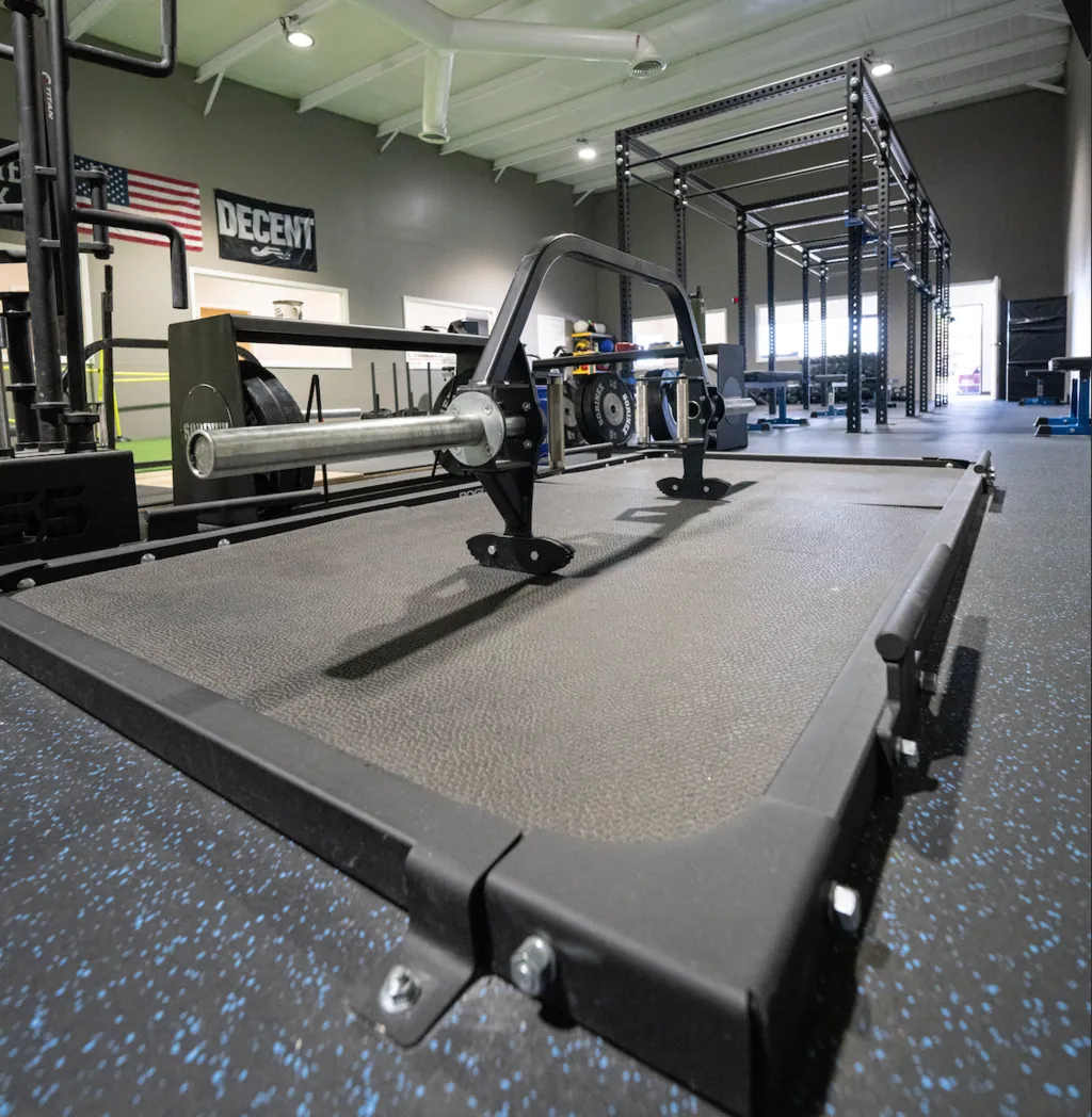 Gym interior with a hex barbell resting on a rubber platform, weight racks, and a pull-up rig in the background.