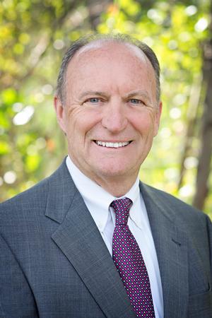 Smiling middle-aged man in suit and polka dot tie with green leafy background.