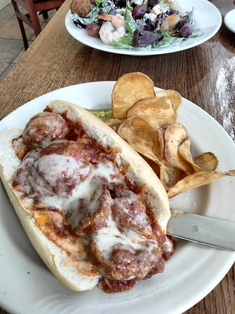 Meatball sub sandwich with melted cheese and marinara sauce served with a side of kettle-cooked potato chips on a white plate.