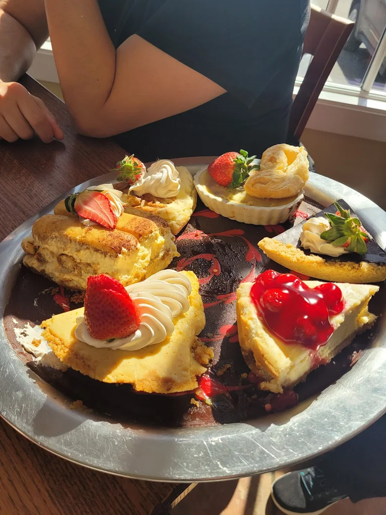 Silver tray with assorted desserts including cheesecake slices topped with strawberries and dollops of cream, tiramisu, and puff pastry, lit by warm sunlight.