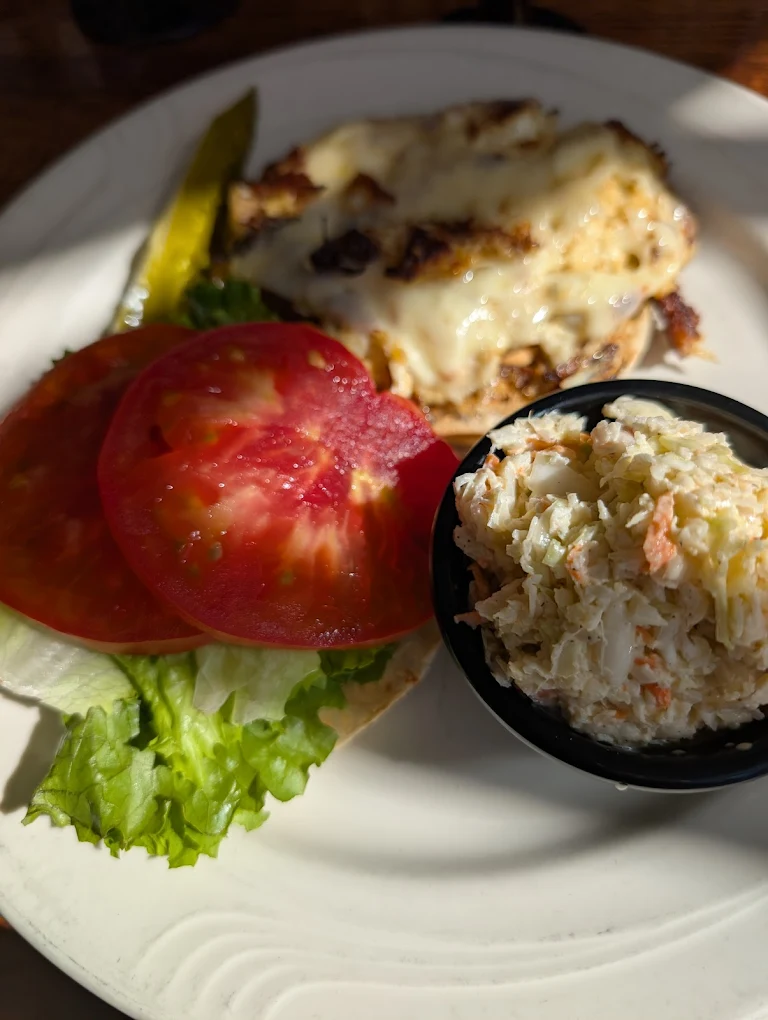 Plate with an open sandwich featuring melted cheese on grilled chicken, lettuce, tomato slices, a pickle spear, and a side of coleslaw in a black cup.