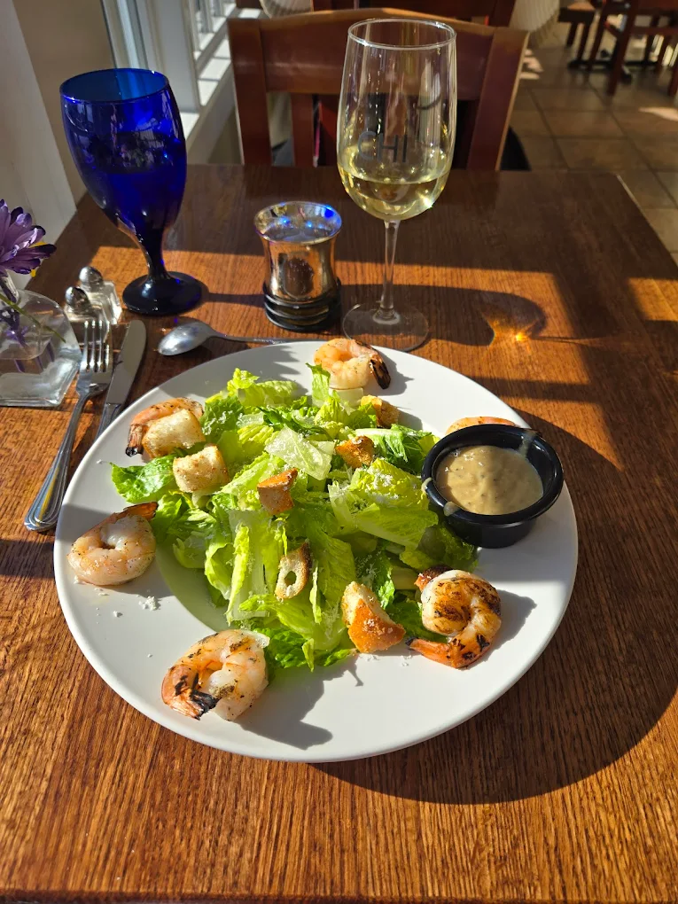 Plate of shrimp Caesar salad with croutons and dressing on a wooden table, accompanied by a glass of white wine and a blue water glass.