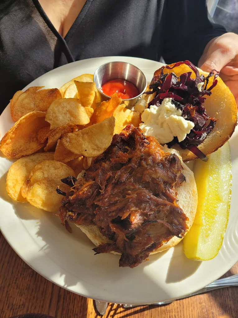 White plate with pulled pork sandwich topped with slaw and cheese, a side of potato chips, ketchup in a metal cup, and a pickle spear.
