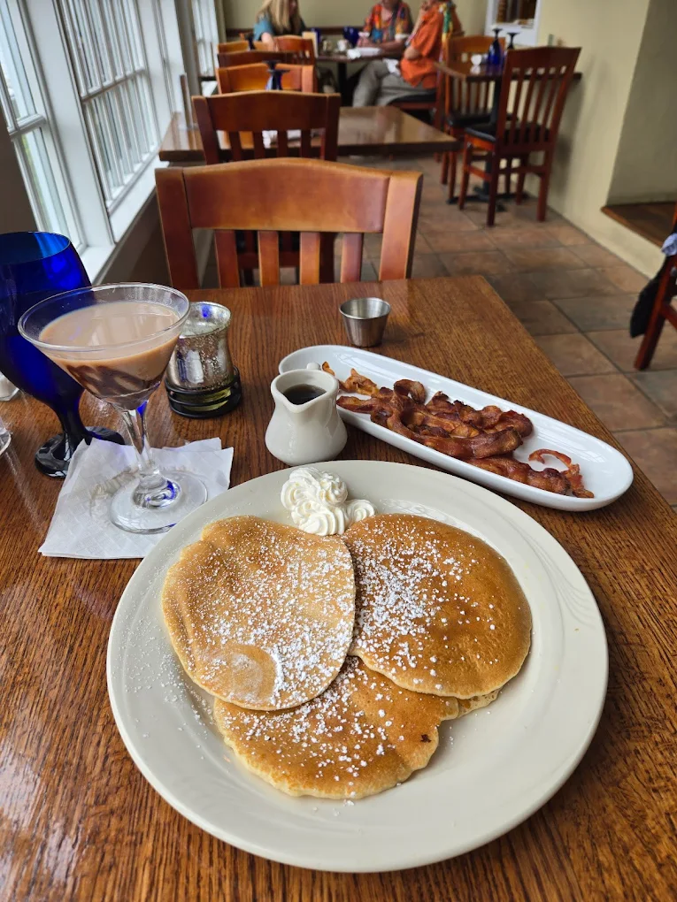 Plate with three powdered sugar-topped pancakes and whipped cream, served with a side of crispy bacon and syrup on a wooden table inside a restaurant.
