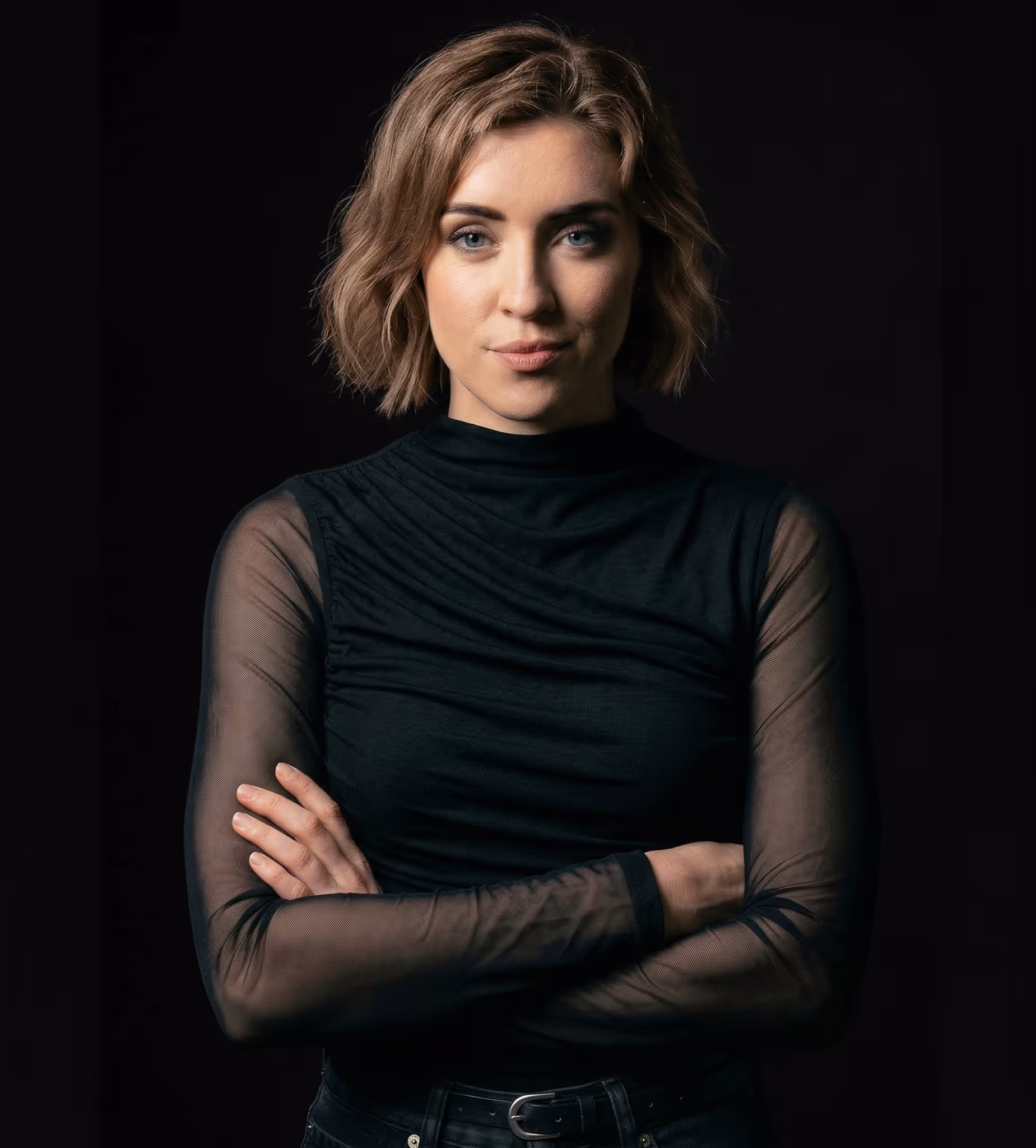 Confident woman with short blonde hair wearing a black sheer-sleeve top and arms crossed.