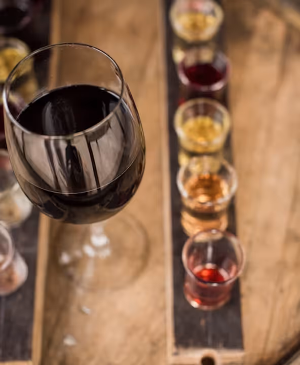 Close-up of a glass of red wine in the foreground with a wooden tray holding multiple small glasses filled with different colored liquids in the background.