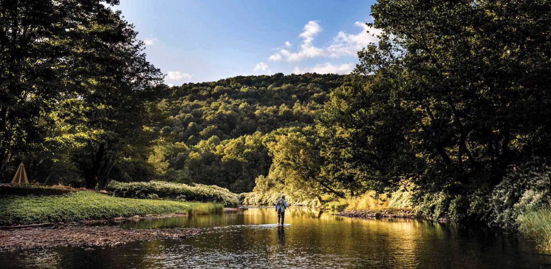 Person fly fishing while standing in a calm river surrounded by dense green trees and hills under a blue sky.