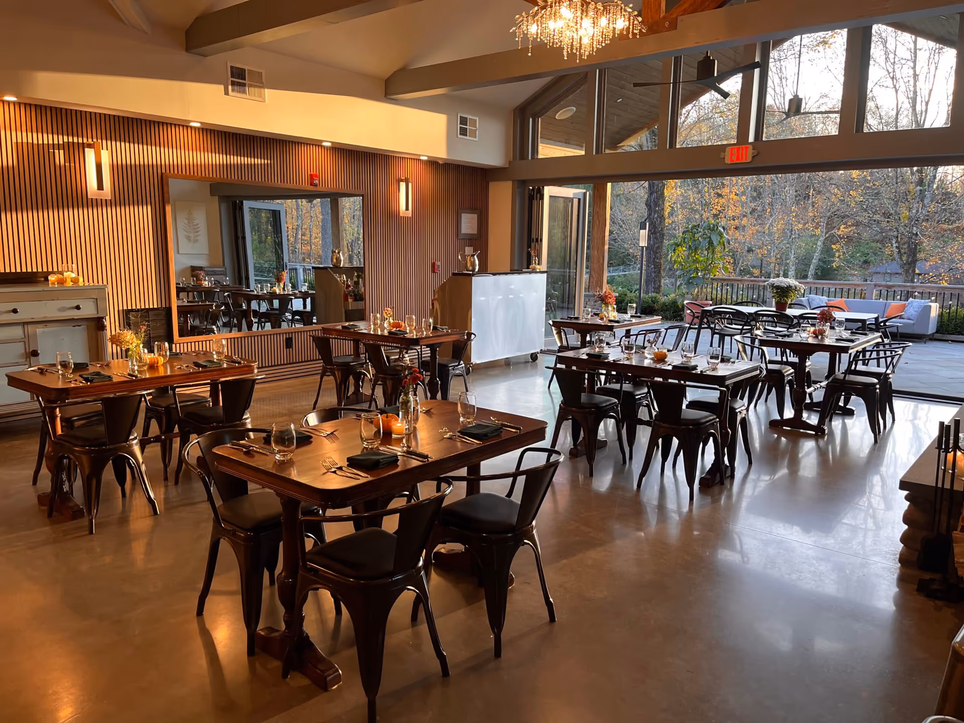 Cozy restaurant interior with wooden tables, black chairs, and autumn decorations near large windows overlooking trees.