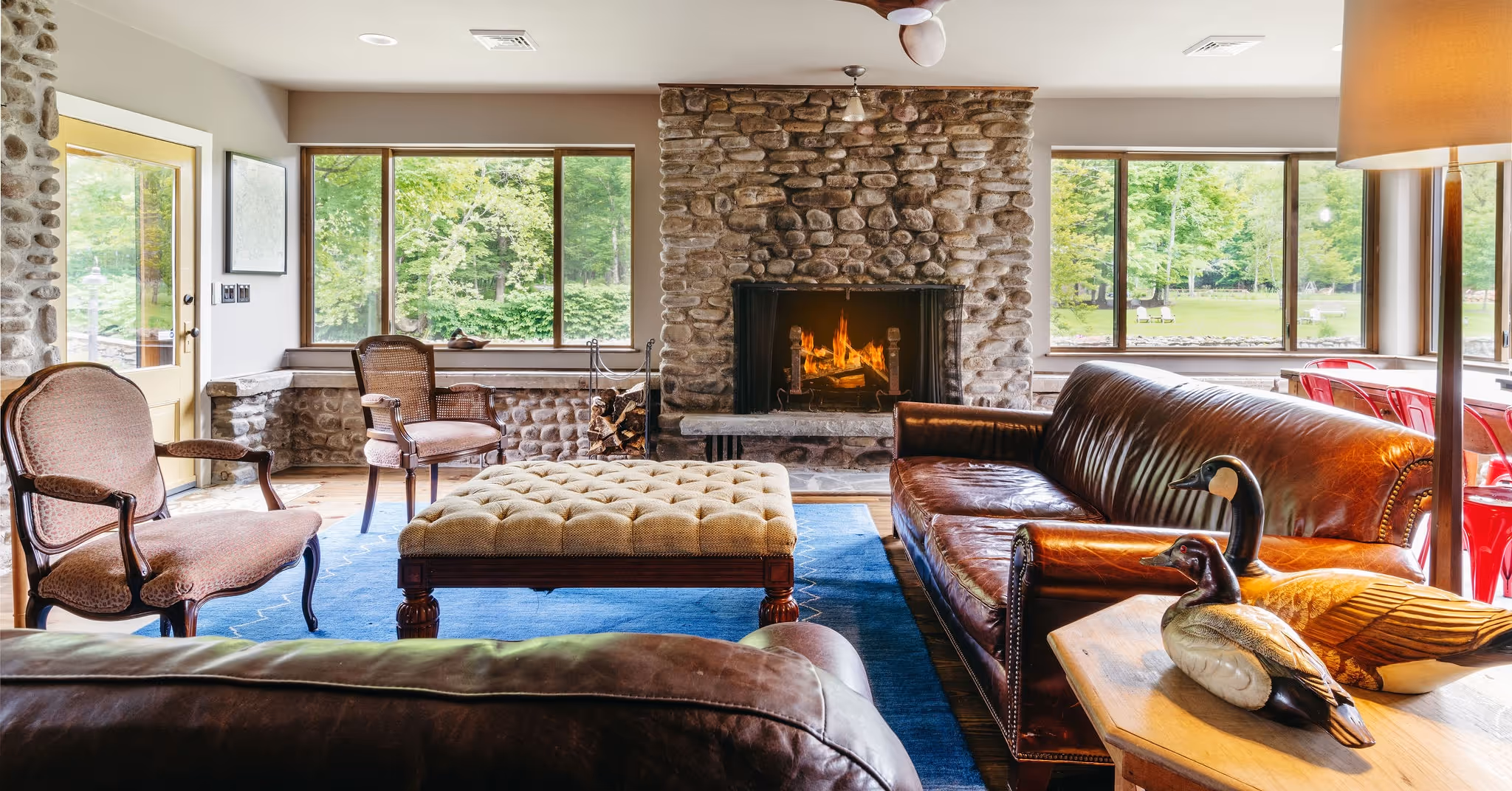 Cozy living room with stone fireplace, leather sofas, wooden duck decorations, and large windows overlooking green trees.