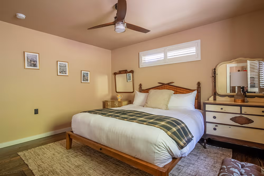 Cozy bedroom with a wooden bed frame, white bedding, green plaid throw, beige walls, ceiling fan, and vintage dresser with a mirror.