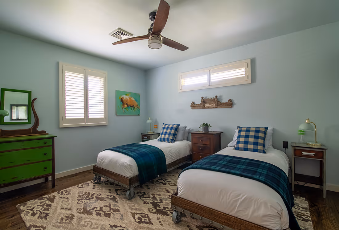 Bedroom with two twin beds featuring white bedding, blue plaid pillows, and blue throws, between two windows with white shutters.