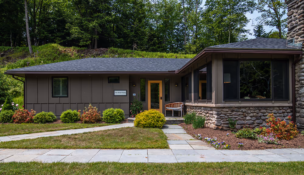 Modern single-story house with a stone foundation, brown vertical siding, a gray shingled roof, a bench near the entrance, and surrounding green landscaping.