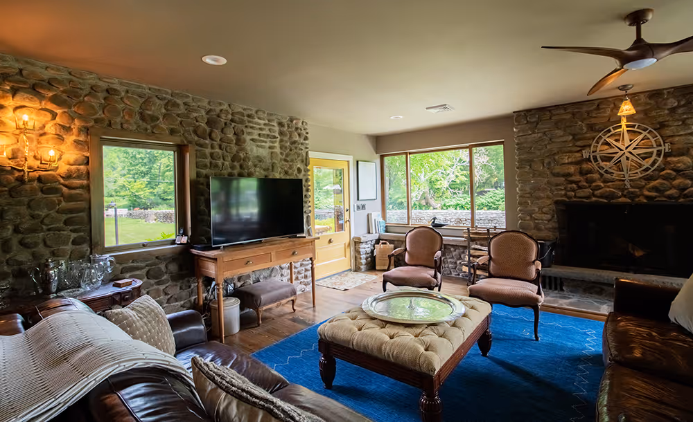 Cozy living room with stone walls, large windows, leather sofas, a tufted ottoman on a blue rug, and a fireplace with a compass wall decoration.