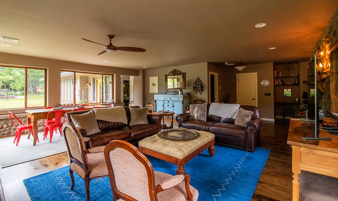 Cozy living room with leather sofas, cushioned ottoman on a blue rug, upholstered chairs, and a dining area with red chairs near large windows.