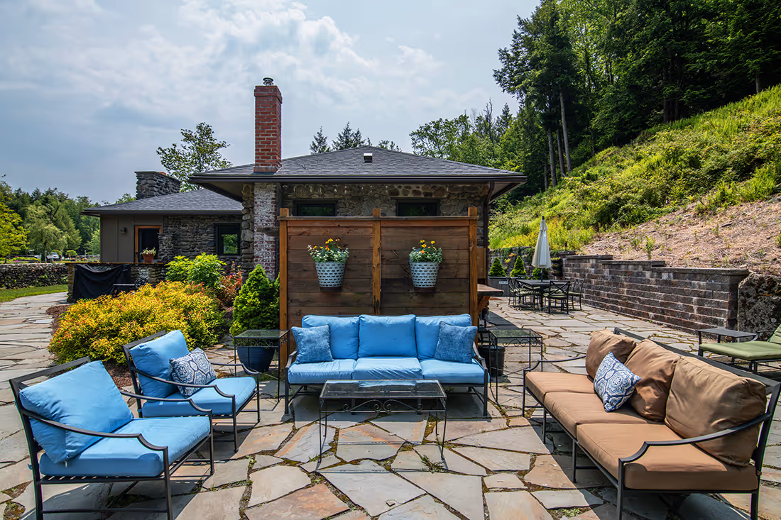 Outdoor patio with blue and brown cushioned seating, a small glass table, and a backdrop of a stone house and greenery.