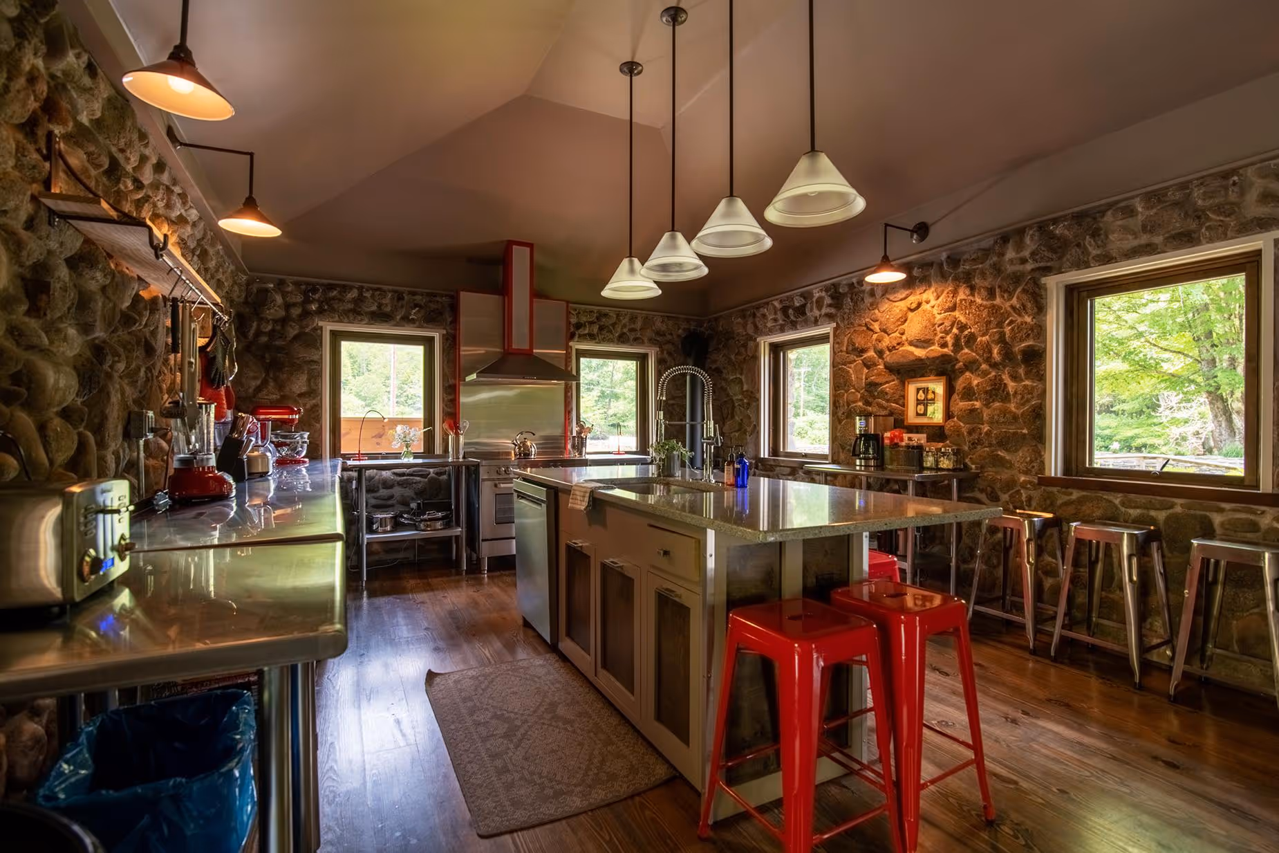 Rustic kitchen with stone walls, hardwood floors, stainless steel appliance counters, an island with red stools, and large windows showing green trees outside.