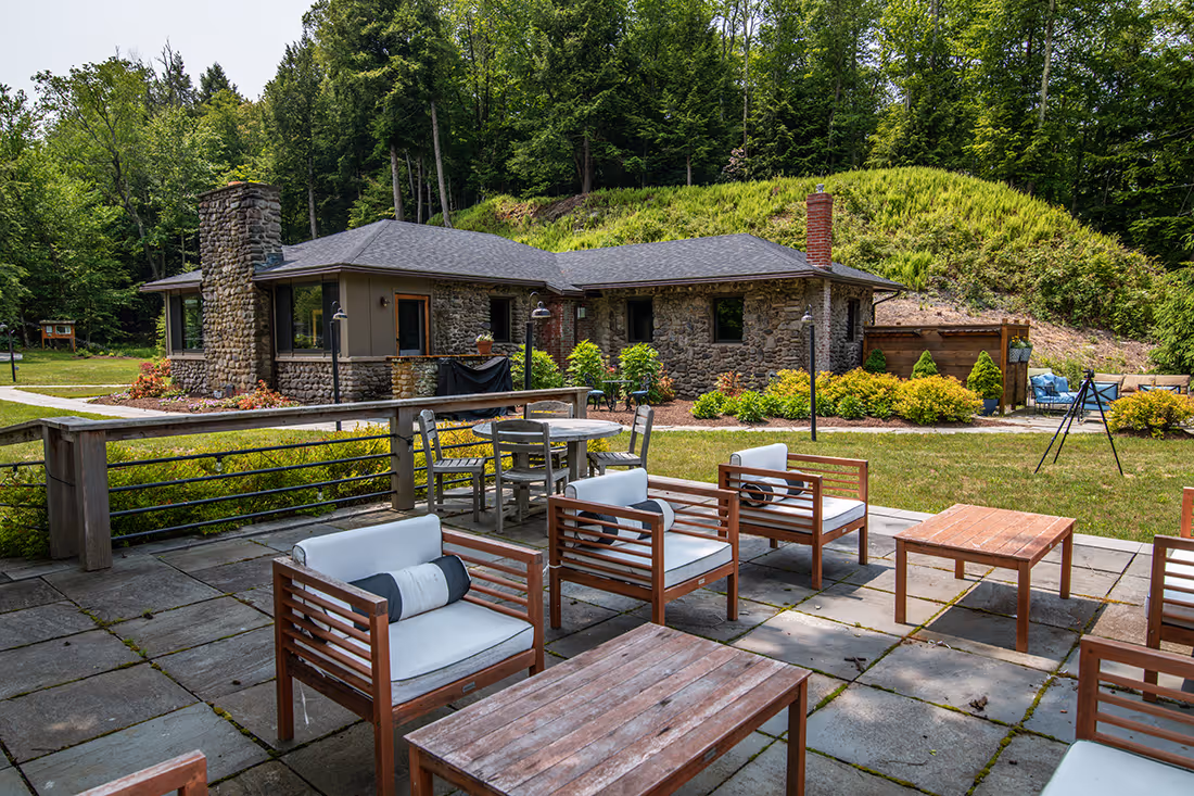 Outdoor patio with wooden chairs and tables overlooking a stone house surrounded by greenery and trees.