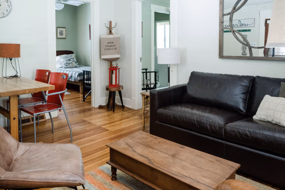 Cozy living room with a black leather couch, wooden coffee table, brown chair, and dining area with red chairs, leading to a bedroom.