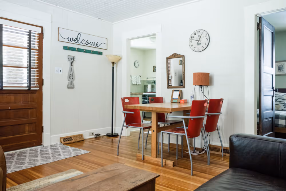 Dining area with a wooden table surrounded by four red chairs in a cozy room with wooden floors, a welcome sign on the wall, a standing lamp, and two open doorways leading to other rooms.