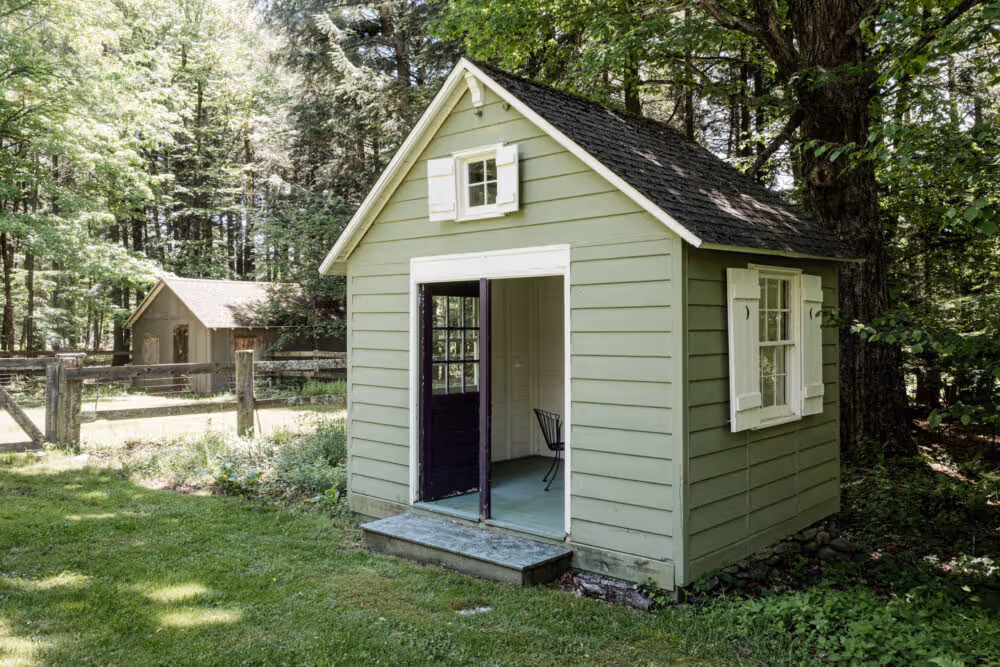 Small green cottage with white shutters and black door, set in a grassy yard surrounded by trees and wooden fence.