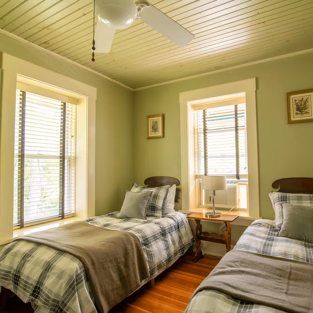 Cozy bedroom with two twin beds dressed in plaid bedding, a wooden nightstand with a lamp between two windows with blinds, and a ceiling fan above.