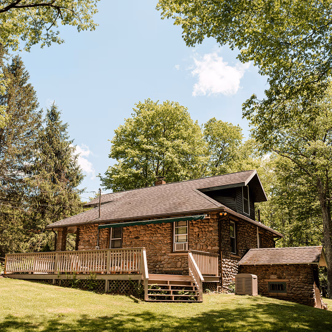 Stone cottage with wooden porch surrounded by green trees under a clear blue sky.