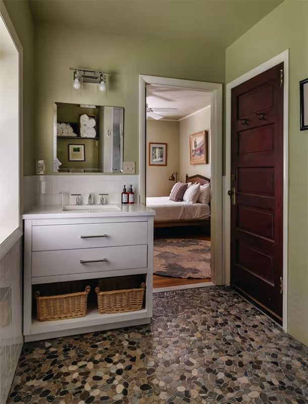 Bathroom with stone pebble floor, white vanity with sink and mirror, green walls, and open doorway leading to a bedroom with a bed and framed pictures.