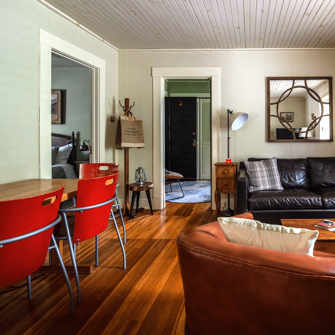 Cozy living room with wooden floor, red dining chairs, leather sofa, floor lamp, decorative mirror, and view into a bedroom and entryway.