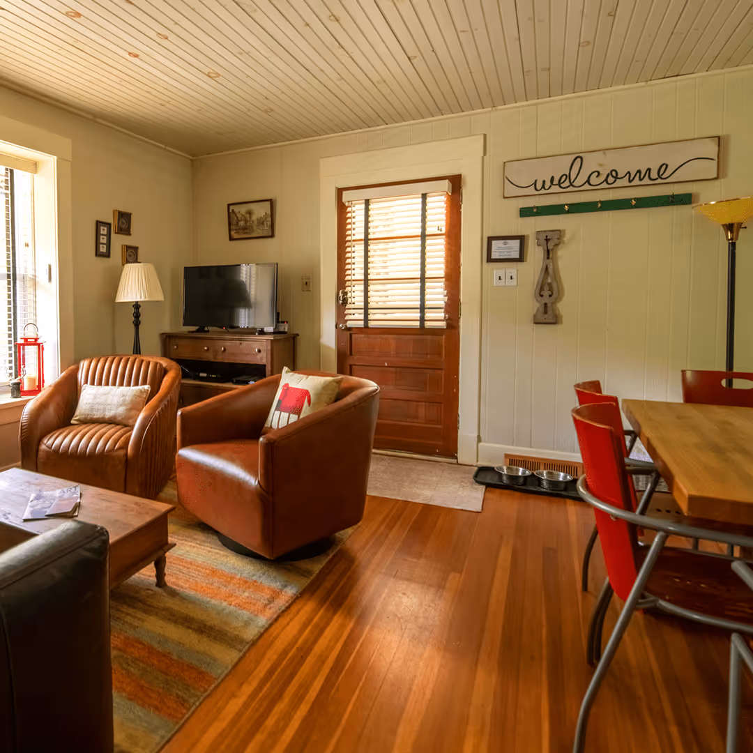Cozy living room with two brown leather armchairs, a wooden coffee table, a TV on a wooden console, and a welcome sign above the wooden door.