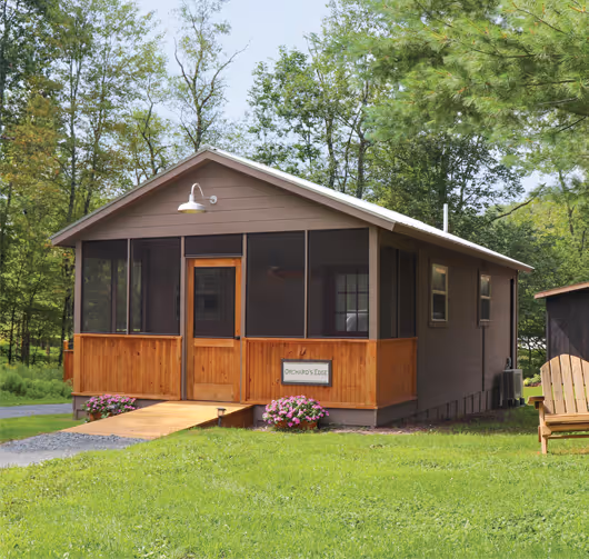 Small cabin with a screened porch and wooden ramp surrounded by green grass and trees.