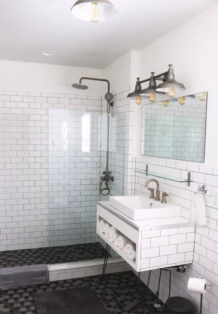 Modern bathroom with white subway tile walls, black hexagonal floor tiles, glass-enclosed shower with rain showerhead, and a white sink with brass faucet and mirror above.