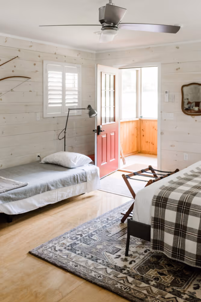 Bright bedroom with a single bed, a bed with plaid blanket, open red door, and wooden paneled walls.
