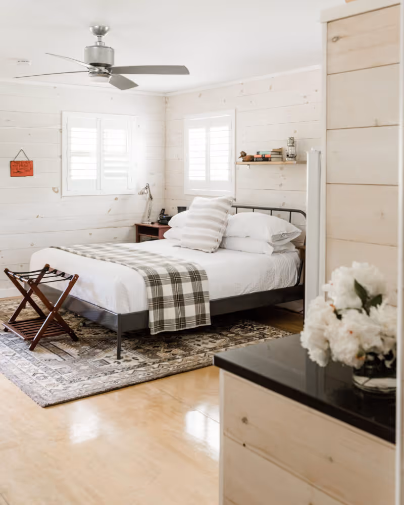 Bright bedroom with white shiplap walls, a metal bed with white linens and plaid throw, wooden luggage rack, and a ceiling fan.