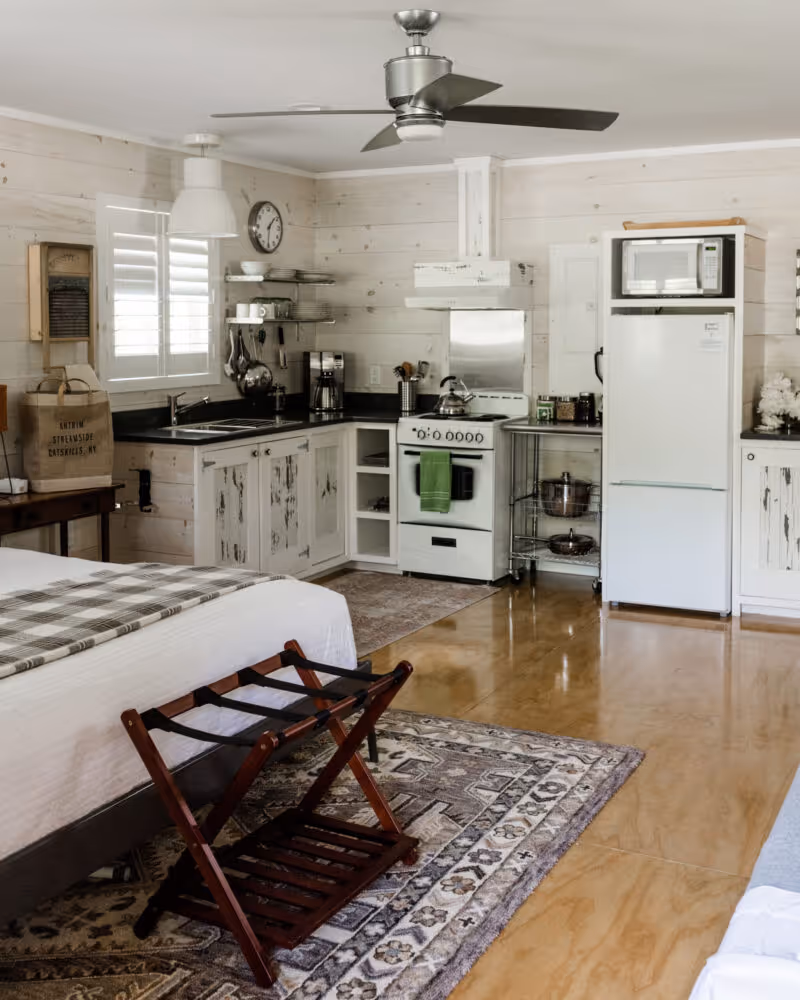 Cozy kitchen corner with white rustic cabinets, stove with green towel, fridge with microwave on top, ceiling fan, and part of a bed with luggage rack in foreground.